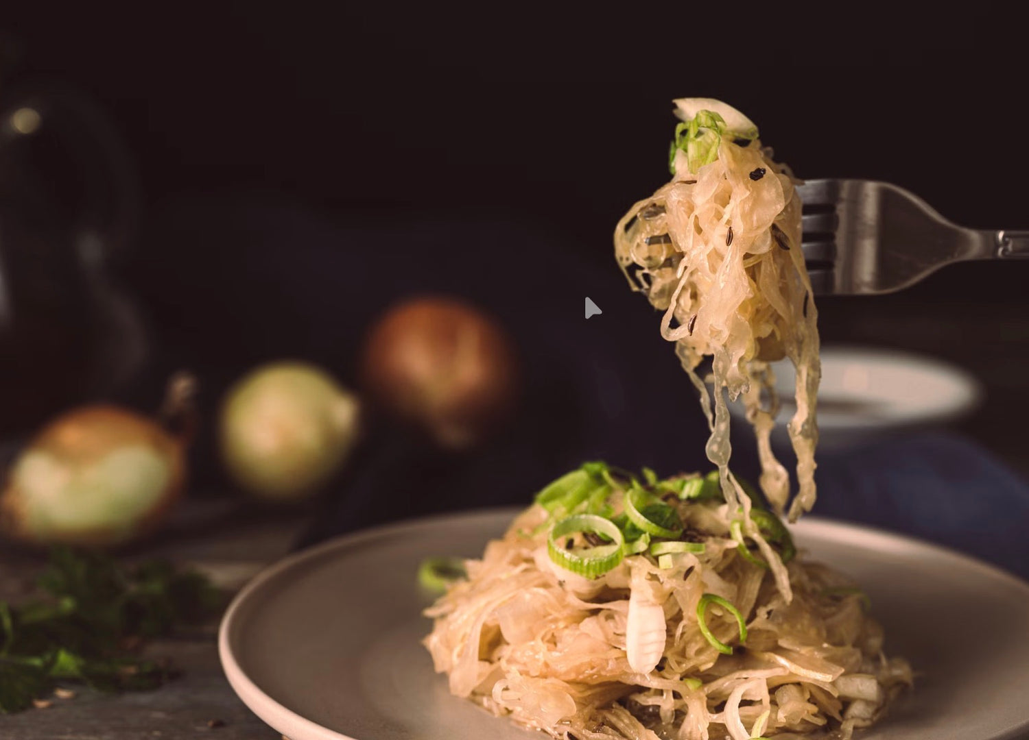 A close-up of a fork lifting a serving of sauerkraut with green onions from a plate, set against a dark, blurred background with onions and herbs.